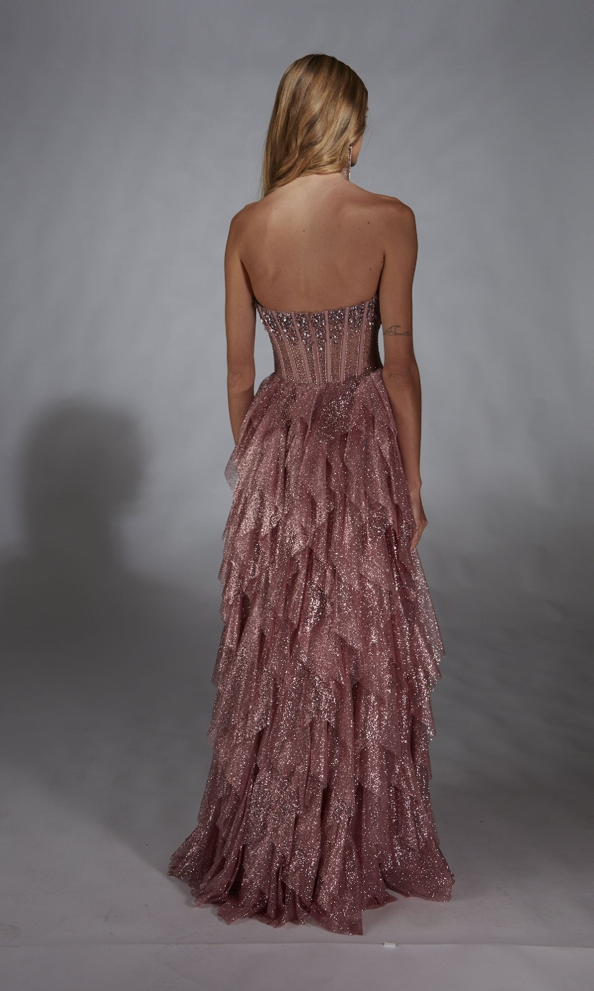 A woman faces away, wearing the Alyce 61762 Strapless Long Glitter A-Prom Dress with a tiered ruffled skirt, set against a plain backdrop.